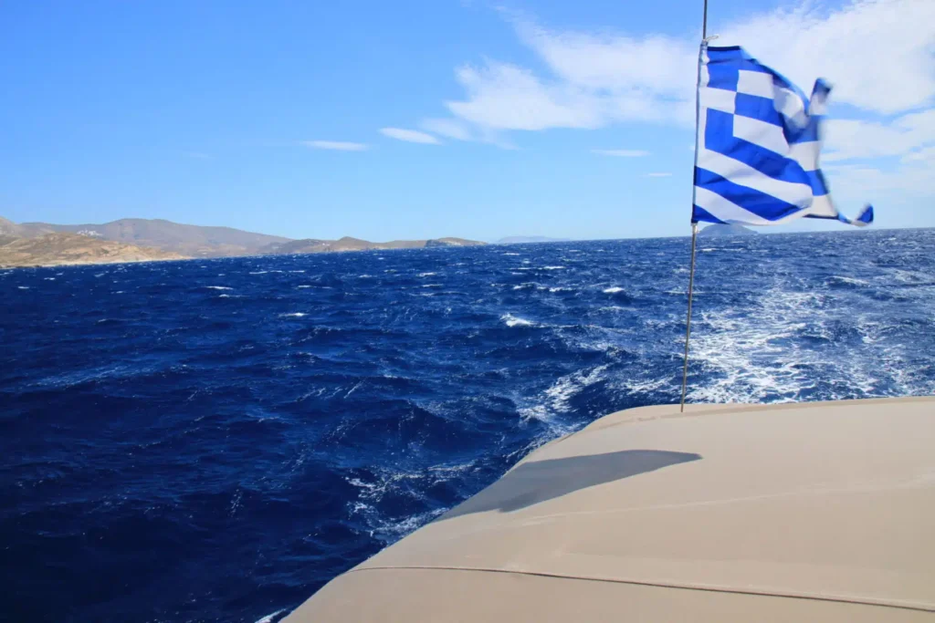 Blick von einer Segelyacht auf das tiefblaue Meer mit einer im Wind wehenden griechischen Flagge und einer bergigen Küste im Hintergrund unter einem klaren Himmel.