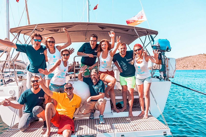 Eine Gruppe fröhlicher Menschen posiert mit aufgeregt erhobenen Händen auf einem Segelboot, trägt passende T-Shirts und genießt einen Segelurlaub mit klarem Himmel und ruhigem blauen Wasser im Hintergrund.