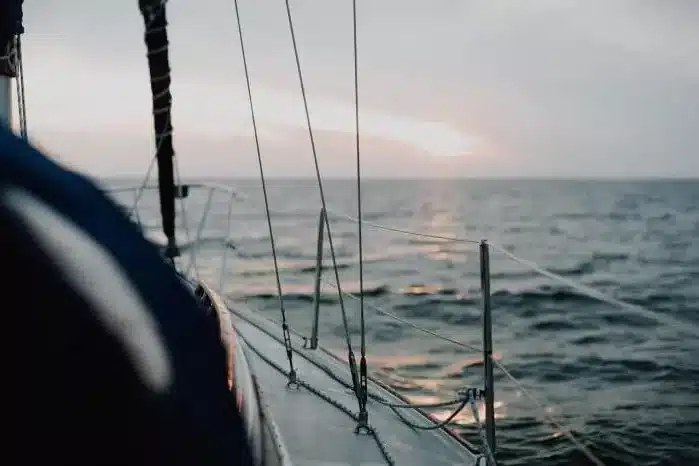 Blick von der Seite einer Segelyacht in der Abenddämmerung, mit Fokus auf die Metalltakelage mit dem Meer und einem sanften Sonnenuntergang im Hintergrund.