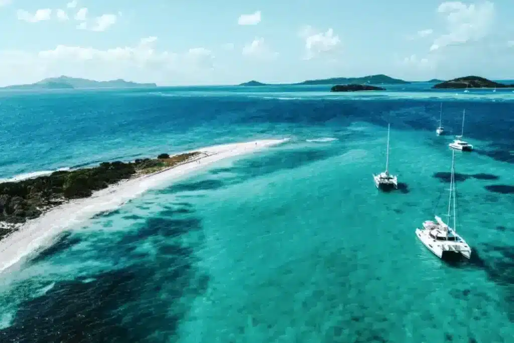 Luftaufnahme eines schmalen Sandstrands mit kristallklarem, türkisfarbenem Wasser, umgeben von mehreren Segelyachten vor einer üppig grünen Insel.