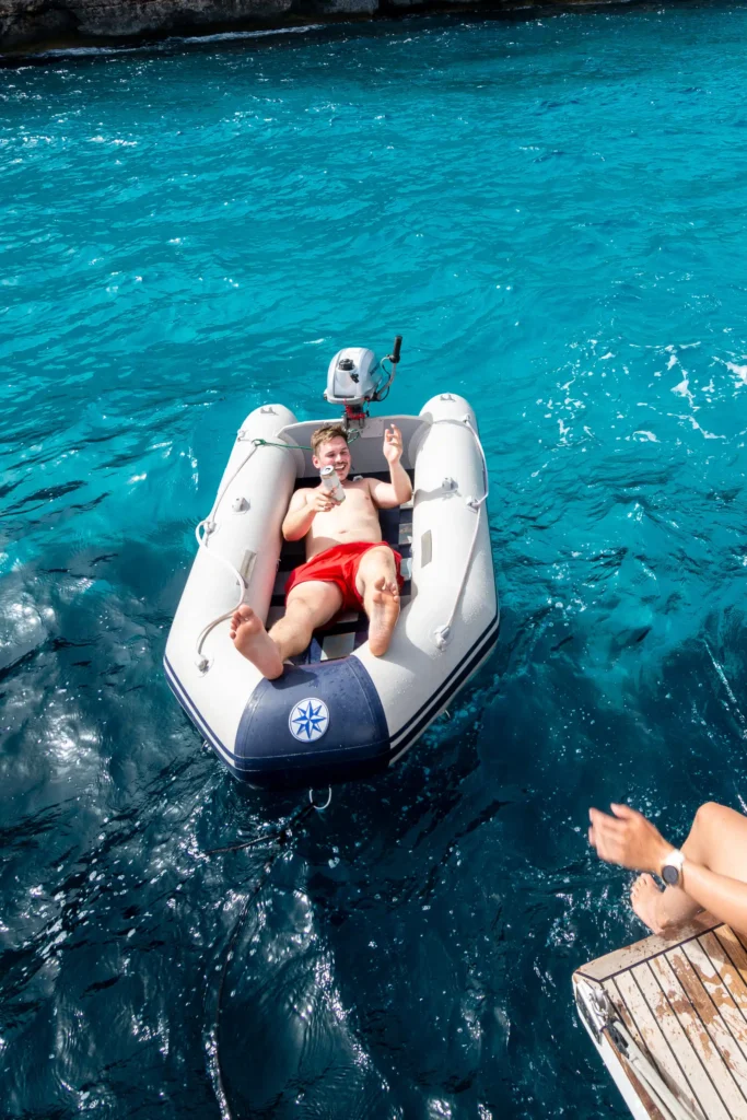 Eine Frau sitzt auf einer Bootsplattform, lässt ihre Beine in Richtung Wasser baumeln und winkt zwei lächelnden Personen in einem kleinen Schlauchboot zu. Sie treiben im hellen Sonnenlicht auf klarem, türkisfarbenem Wasser in der Nähe einer Felsklippe. Auf der Klippe wachsen Bäume und Sträucher – eine idyllische Kulisse für ihr Segeltörn-Abenteuer.