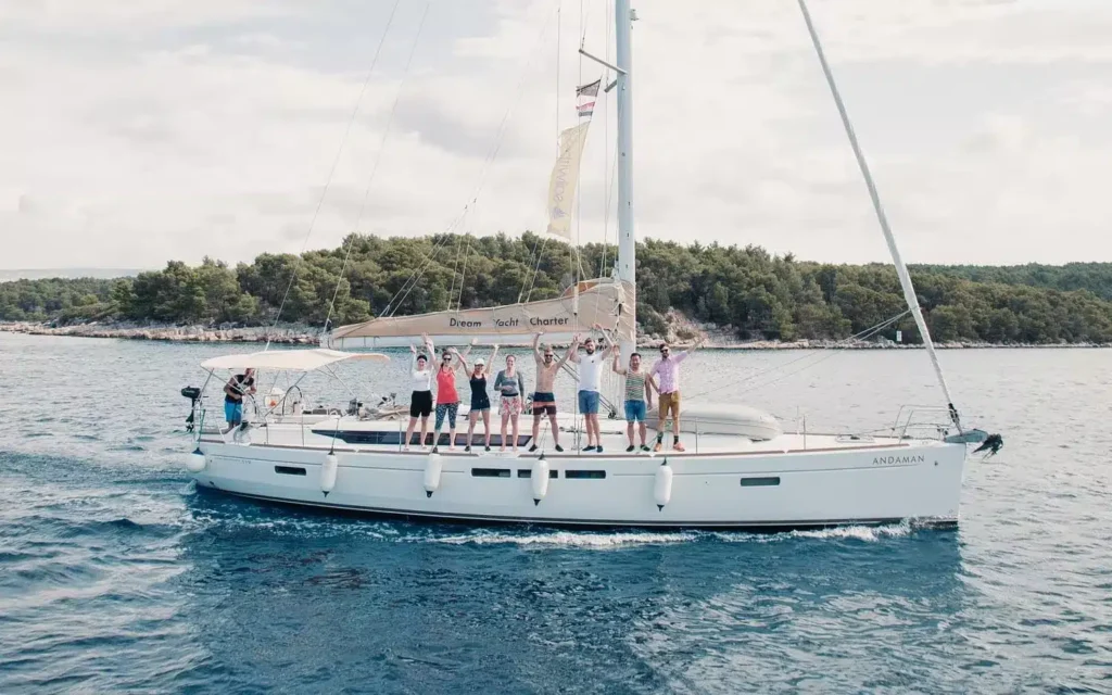 Eine Gruppe von Menschen winkt fröhlich vom Deck einer weißen Segelyacht, die während ihres Segeltörns unter einem teilweise bewölkten Himmel in der Nähe einer bewaldeten Küste vor Anker liegt.