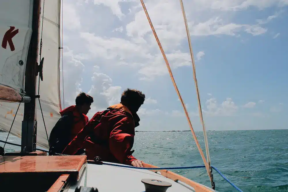 Zwei Menschen in roten Jacken segeln an einem sonnigen Tag mit klarem blauen Himmel und ruhiger See ein kleines Boot.