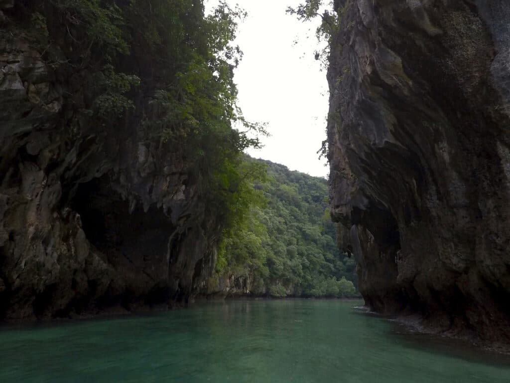 Ein ruhiger Fluss, flankiert von hoch aufragenden, schroffen Klippen, die unter einem bewölkten Himmel mit dichter grüner Vegetation bedeckt sind. Das Wasser ist ruhig und hat einen klaren blaugrünen Farbton, der an die friedliche Fahrt einer Segelyacht erinnert.