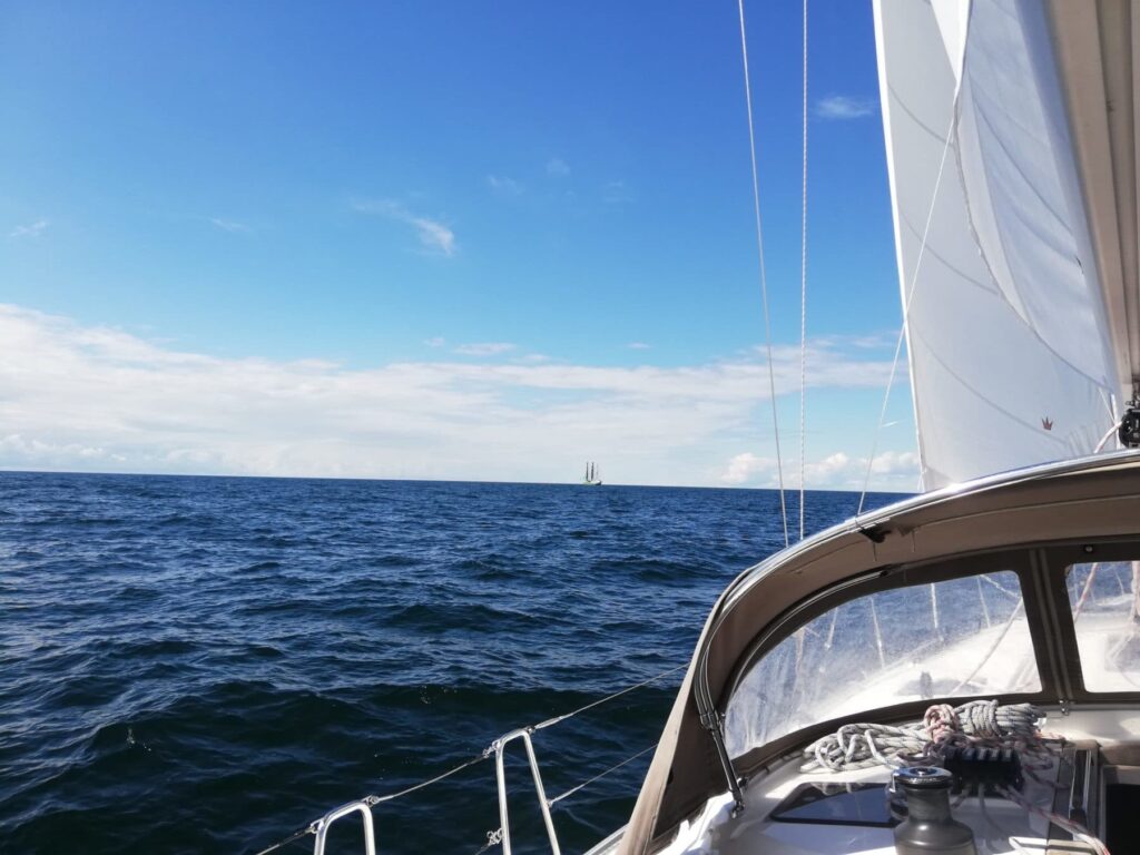 Ein Blick aus dem Cockpit einer Segelyacht. Zu sehen sind ein weißes Segel und die Takelage des Bootes, vor uns das offene Meer und in der Ferne ein weiteres Segelboot unter einem klaren blauen Himmel.
