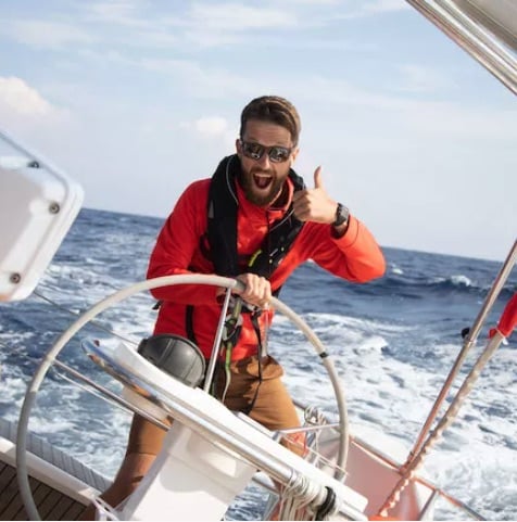 Ein Mann in einer roten Jacke und mit Sonnenbrille steuert einen Katamaran und drückt mit einem erhobenen Daumen seine Begeisterung an einem sonnigen Tag vor der Kulisse des strahlend blauen Ozeans und der weißen Wellen aus.